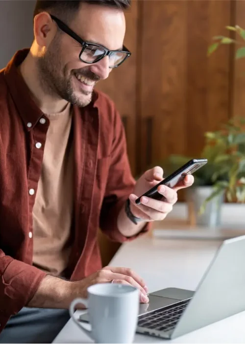 Smiling man using phone and laptop for website design services by Miami Epic Tees
